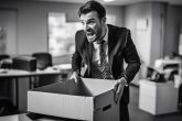 Image en noir et blanc d'un employé de bureau grimaçant en essayant de soulever une lourde boîte, indiquant une douleur due à une maladie discale dégénérative, avec un bureau flou en arrière-plan.