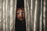 A terrified man peering through split-curtains at a jovial-looking dwarf with dramatic and atmospheric lighting focus.