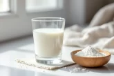 Un verre de lait crémeux posé à côté d’une poudre blanche sur une surface neutre.