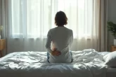 Une personne assise au bord d’un lit tient sa main sur le bas du dos, un document médical à côté d’elle, dans une chambre paisible éclairée par la lumière du matin.