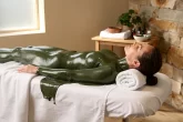 Personne allongée sur un lit de spa, recouverte de boue thérapeutique verte dans une salle apaisante à la lumière naturelle.