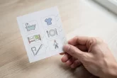Une main tient délicatement une liste de contrôle ornée de six icônes représentant des activités quotidiennes, posée sur une table en bois sous une lumière naturelle douce.