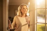 Une femme d’âge mûr se tient devant une porte ouverte baignée de lumière matinale, le regard tourné vers l’extérieur avec espoir.