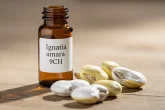 Un flacon ambré de remède homéopathique Ignatia amara 9CH est posé à côté de fèves blanches et jaunes de Saint-Ignace sur une table en bois éclairée doucement.