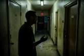 Un homme seul se tient devant une porte d’appartement faiblement éclairée dans un couloir nocturne, partiellement illuminé par une lumière vacillante.