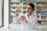 Un pharmacien en blouse blanche examine un flacon d’eau du robinet devant un comptoir ordonné, avec des rayons de médicaments en arrière-plan et une lumière du jour douce.