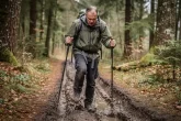 Un randonneur d’âge mûr s’arrête sur un sentier boueux de forêt, l’air frustré, son soulier coincé dans la boue et tenant des bâtons de marche nordique.