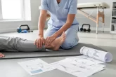 Un thérapeute aide un patient à réaliser des exercices de rééducation du pied sur un tapis, près de feuilles de protocole et sous une lumière matinale douce.